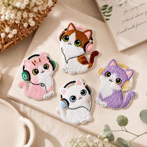 May include: Embroidered patches featuring cartoon cats wearing headphones. The patches are in various colors, including pink, white, brown, and purple. Each cat has large eyes and a cute expression. The patches are likely for clothing or accessories.