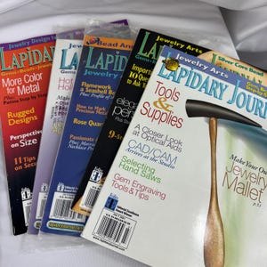Può includere: Una pila di riviste di gioielleria, tra cui "Lapidary Journal" e "Jewelry Arts." Le copertine presentano design colorati e articoli su strumenti, forniture e tecniche per la creazione di gioielli. Le riviste sono impilate.