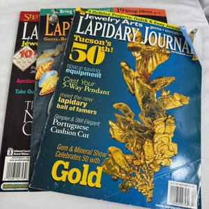 Könnte beinhalten: Drei Ausgaben des Lapidary Journal Magazins sind auf einer weißen Oberfläche gestapelt. Die Dezemberausgabe 2003 zeigt eine goldfarbene Mineralformation auf blauem Hintergrund. Andere Ausgaben haben Texte und Bilder zu Schmuck und Edelsteinen.