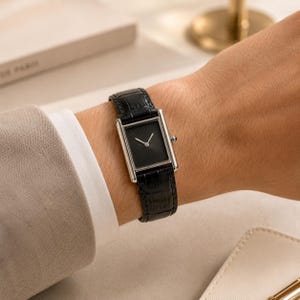 May include: A rectangular wristwatch with a silver-toned case and a black dial featuring a sunray pattern. The watch has a black leather strap with a crocodile-embossed texture and a small silver crown on the side for adjusting the time settings.