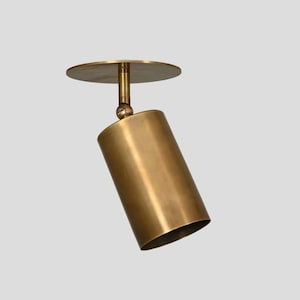 Könnte beinhalten: Ein zylindrischer Strahler in Messingfarbe mit einer kreisförmigen Deckenhalterung. Der Strahler ist mit einem Kugelgelenk verstellbar und nach unten geneigt. Die Leuchte hat ein modernes, minimalistisches Design.