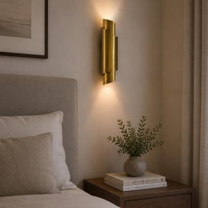 Könnte beinhalten: Eine goldfarbene Wandleuchte mit zylindrischem Design an einer hellen Wand. Die Leuchte strahlt warmes Licht aus. Ein Nachttisch mit Büchern und einer Vase mit Grünzeug steht neben einem Bett.