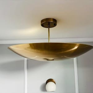 Könnte beinhalten: Eine messingfarbene Deckenleuchte aus Metall mit einem breiten, flachen, schalenförmigen Schirm, der an einer dünnen Mittelstange von einem runden Baldachin hängt. Unter der Hauptleuchte ist eine kleine, kugelförmige Wandleuchte aus weißem Glas zu sehen.