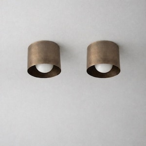 Könnte beinhalten: Zwei zylindrische Deckenleuchten mit gebürstetem Antikmessing-Finish. Jede Leuchte verfügt über einen breiten Metallschirm mit offener Unterseite, in dem sich eine einzelne, kleine, kugelförmige weiße Glühbirne befindet.