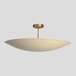 May include: A ceiling light fitting featuring a wide, shallow, off-white bowl-shaped shade suspended by a thin, vertical brass-coloured rod connected to a small, circular brass-coloured canopy mount at the top centre of the fitting.