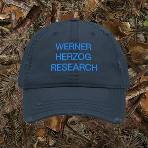 May include: A dark navy blue distressed baseball cap featuring the embroidered text WERNER HERZOG RESEARCH in bright blue thread. The cap has a curved brim and a worn, frayed appearance along the edges of the crown and the bill of the hat.