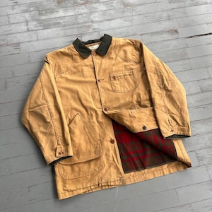 May include: A tan canvas jacket featuring a dark green corduroy collar, a button-front closure, and two large front pockets. The interior lining is visible, showcasing a red and green plaid pattern. The jacket has long sleeves with buttoned cuffs.