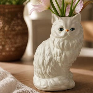 Peut inclure: Un vase en céramique blanche en forme de chat à poils longs avec une texture de fourrure détaillée, des oreilles pointues et des yeux bleus. Le chat a un petit nez orange et une large ouverture sur le dessus de la tête, contenant des tulipes.