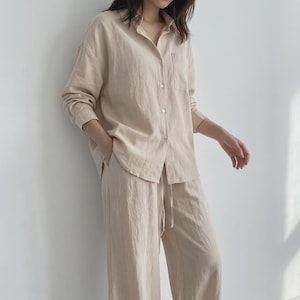 May include: A beige linen pajama set. The long-sleeved button-down shirt has a chest pocket. The pants have a drawstring waist. The fabric appears lightweight and breathable, suitable for sleepwear or loungewear.