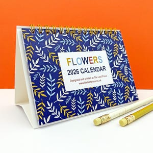 Puede incluir: Un calendario de escritorio de 2026 con un diseño floral. El calendario presenta un fondo azul con patrones de hojas blancas, amarillas y azul claro. El texto "FLOWERS 2026 CALENDAR" se muestra en la parte delantera. Dos lápices están en primer plano.