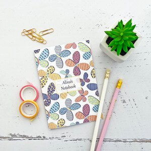 May include: A white notebook with a colourful floral pattern and the text "Alina's Notebook" on the cover. The notebook is on a white wooden surface with two pencils and a roll of washi tape.