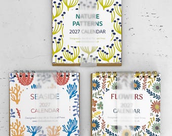 May include: Three square 2027 calendars with wire-o binding. The top calendar features a green and blue nature pattern. The bottom left calendar shows a red, orange, and blue seaside theme. The bottom right calendar displays a floral pattern.