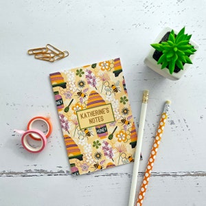 May include: A yellow and white notebook with a bee and honeycomb design. The notebook has the text "Katherine's Notes" on the front cover.