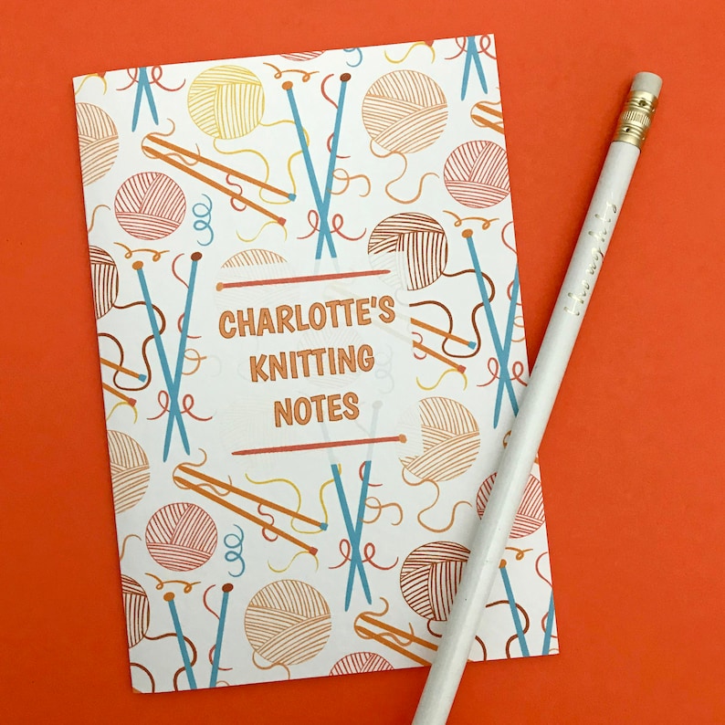 May include: A white notebook with a colorful knitting themed pattern. The notebook has the text "Charlotte's Knitting Notes" on the front cover.