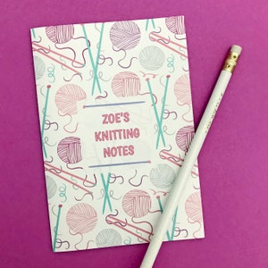 May include: A white notebook with a pink and blue knitting themed pattern. The notebook has the text "ZOE'S KNITTING NOTES" on the front.