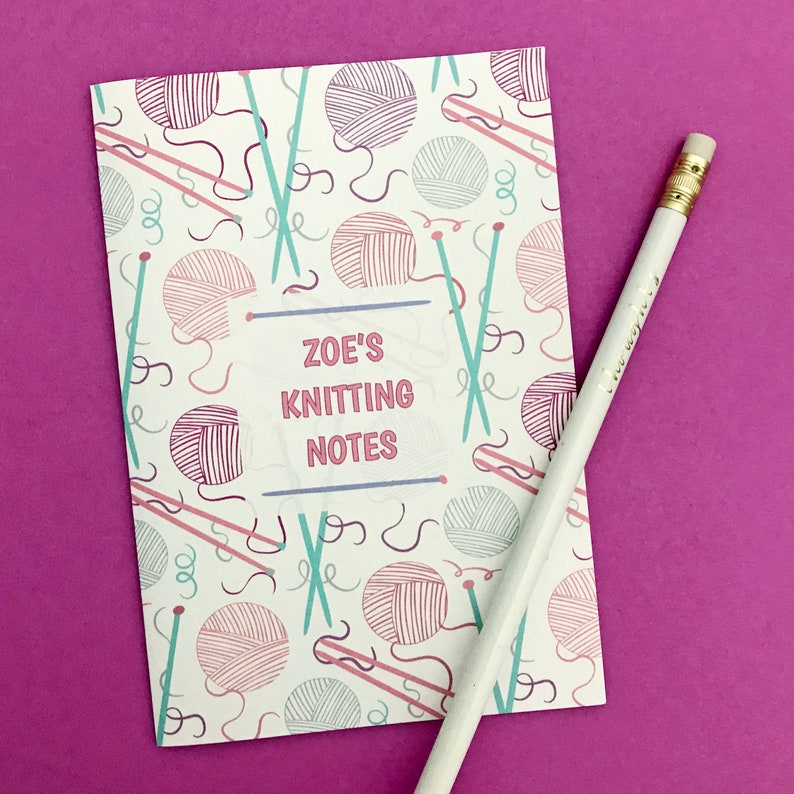 May include: A white notebook with a pink and blue knitting themed pattern. The notebook has the text "ZOE'S KNITTING NOTES" on the front.