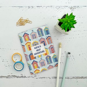 Può includere: Un piccolo quaderno blu e bianco con un motivo colorato di casette sulla spiaggia. Il quaderno ha il testo "Katie's Next Adventure" sulla copertina.