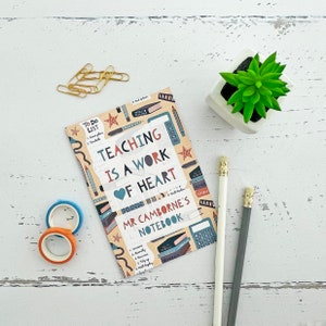 May include: A colourful notebook with the text "Teaching is a work of heart" and "Mr. Camborne's Notebook". The cover features illustrations of school supplies, including a heart, stars, and a calculator.