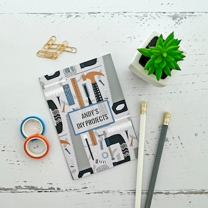 May include: A grey and white notebook with a colourful pattern of tools and the text "ANDY'S DIY PROJECTS". The notebook is on a white wooden surface with two small blue and orange tape dispensers and a small potted succulent plant.