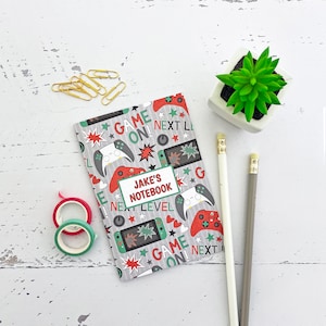 May include: A gray and white notebook with a red, green, and white gaming theme. The notebook has the text "JAKE'S NOTEBOOK" on the front cover. The notebook is surrounded by pencils, paper clips, and washi tape.