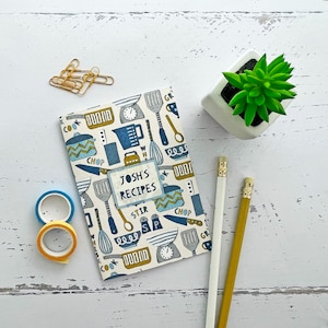 Puede incluir: Un libro de recetas azul y blanco con una ilustración colorida de utensilios de cocina y el texto "Josh's Recipes".