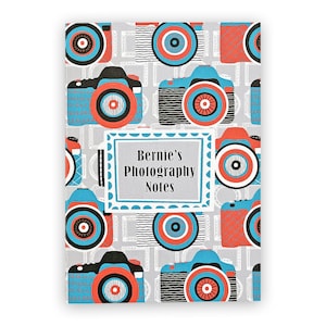 May include: A grey and white notebook with a blue and red camera pattern. The cover has the text "Bernie's Photography Notes" in a white box.