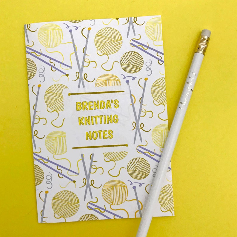 May include: A white notebook with a yellow and gold knitting themed pattern. The notebook has the text "Brenda's Knitting Notes" on the front.