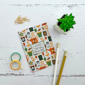Op de afbeelding: Een kleurrijk tuinplanningsnotitieboek met een groen, bruin en geel bloemenontwerp. De omslag heeft de tekst "Alex's Garden Planning".