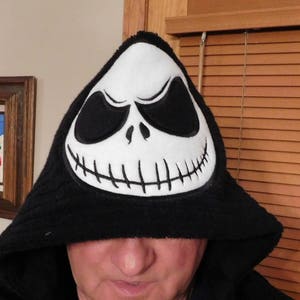 May include: A black hooded sweatshirt with a white Jack Skellington face on the hood. The hood is pulled up over the person's head, covering their eyes.