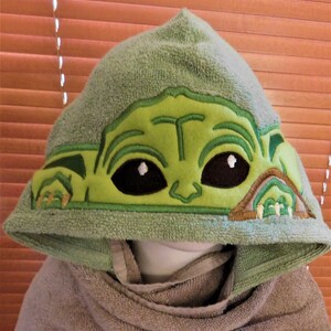 May include: Green hooded towel with an embroidered image of a green alien character with large eyes. The character is wearing a brown ear-like accessory.
