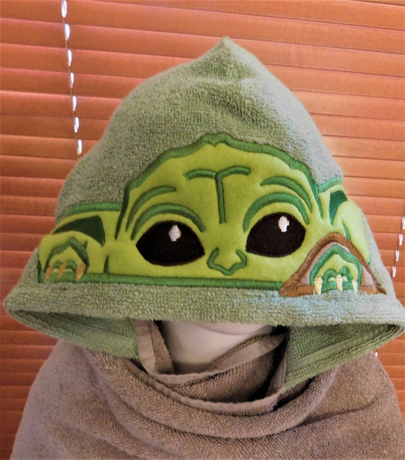 May include: Green hooded towel with an embroidered image of a green alien character with large eyes. The character is wearing a brown ear-like accessory.