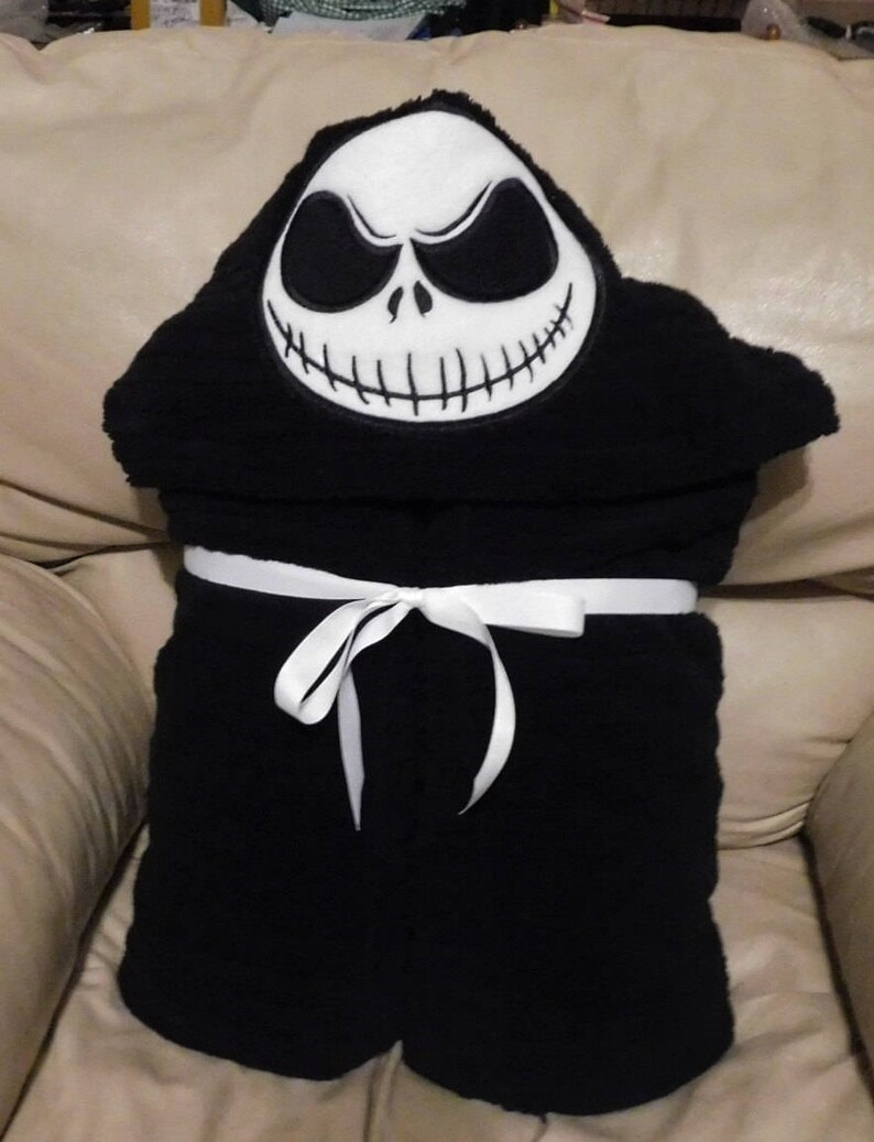 May include: Black hooded towel with a white embroidered Jack Skellington face. The towel has a white ribbon tied around it.