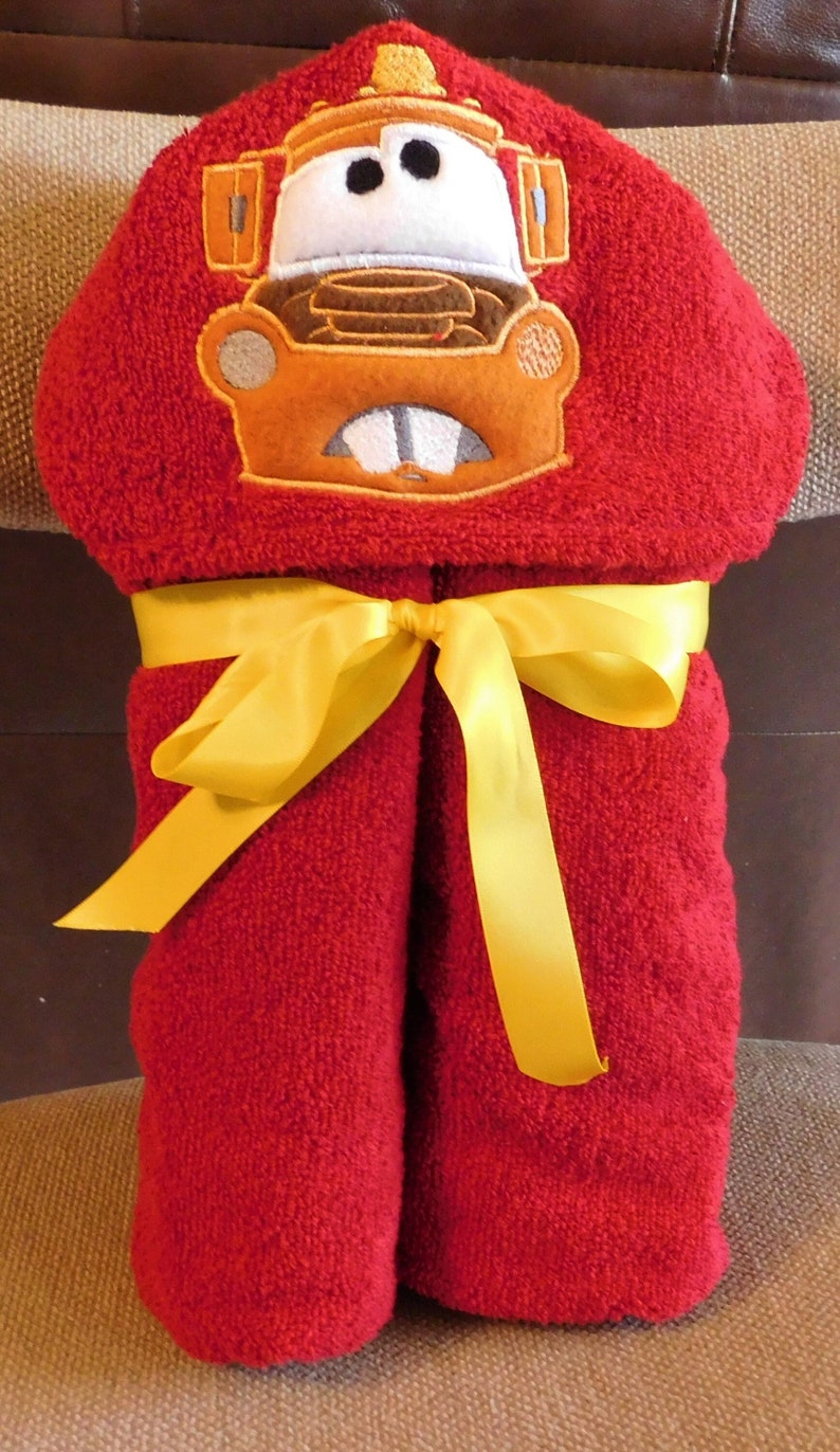 Puede incluir: Toalla con capucha roja con una cinta amarilla. La capucha tiene una imagen bordada de un cami&oacute;n de bomberos de dibujos animados con una cara blanca y ojos negros.