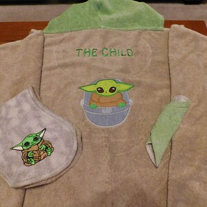 May include: A brown hooded towel with green trim. The towel has an embroidered image of a green alien character, "The Child", from the Star Wars series, on the front. The towel also has a smaller embroidered image of the character on a gray bib.
