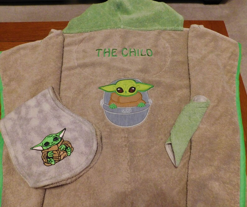 May include: A brown hooded towel with green trim. The towel has an embroidered image of a green alien character, "The Child", from the Star Wars series, on the front. The towel also has a smaller embroidered image of the character on a gray bib.