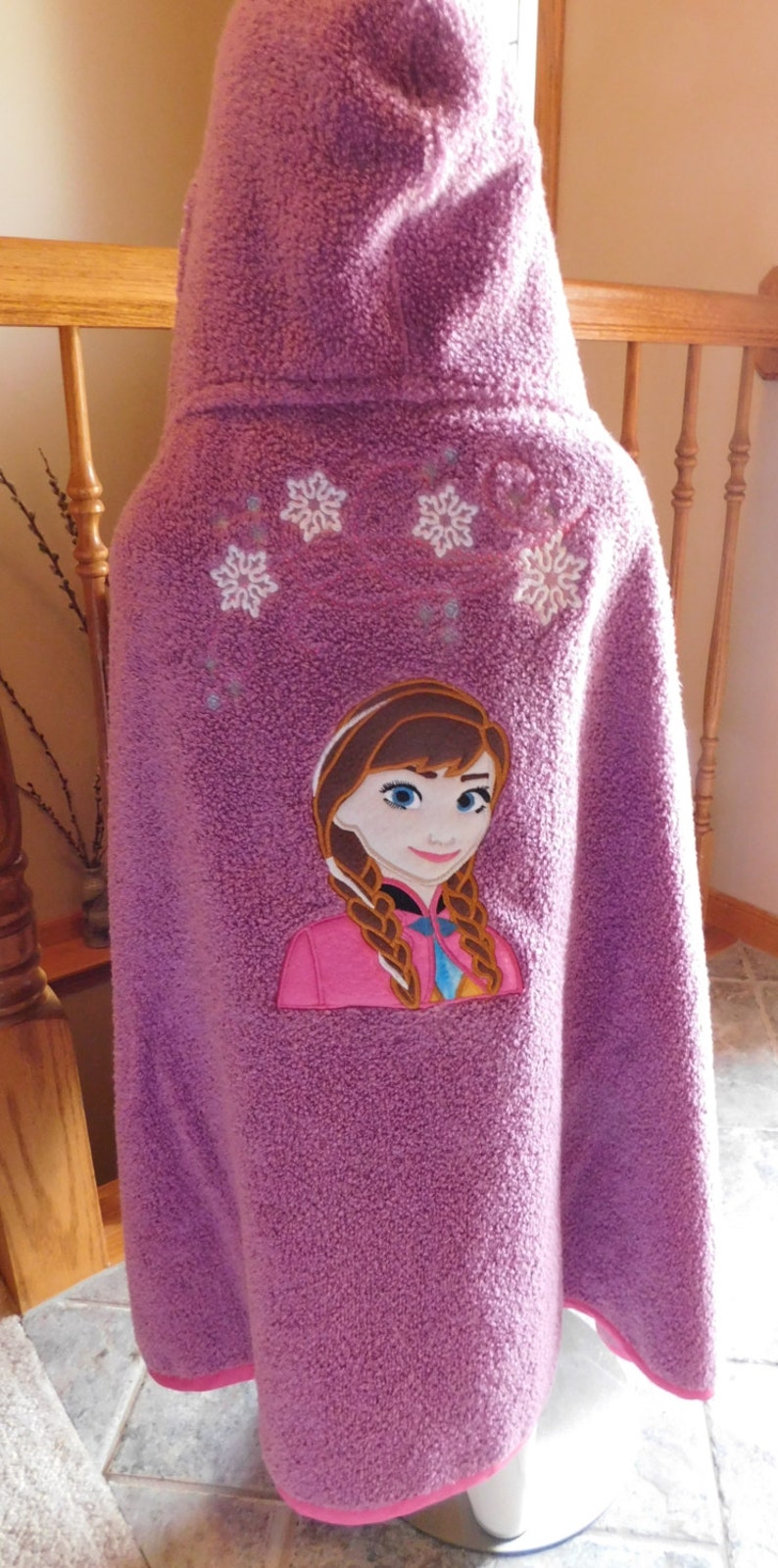May include: Purple hooded towel with an embroidered image of Anna from Frozen. The towel has white snowflakes embroidered on the hood.