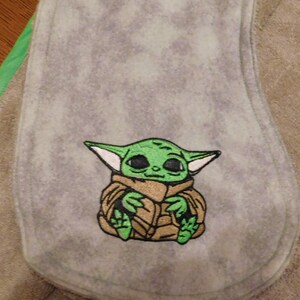 May include: A gray fleece baby burp cloth with an embroidered green and brown image of a character from the Star Wars franchise, The Mandalorian.