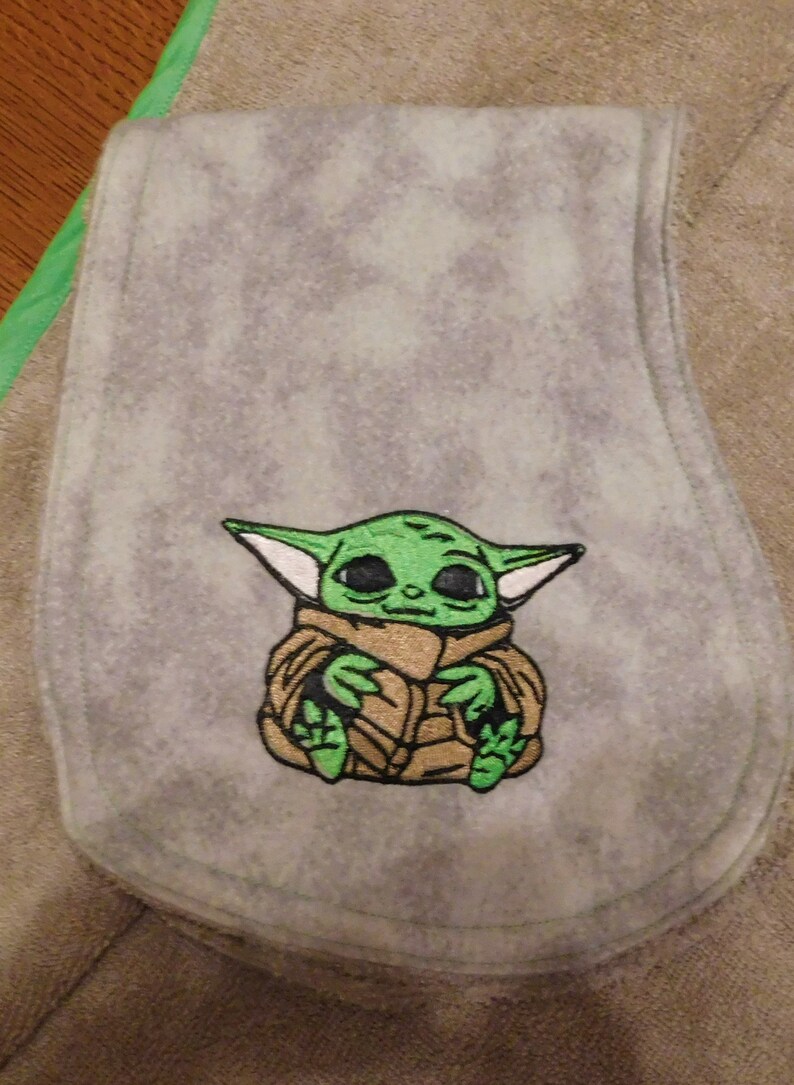 May include: A gray fleece baby burp cloth with an embroidered green and brown image of a character from the Star Wars franchise, The Mandalorian.