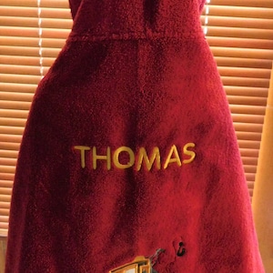 Puede incluir: Toalla con capucha roja con el nombre "THOMAS" bordado en amarillo. Un cami&oacute;n de remolque amarillo con una cara sonriente est&aacute; bordado en la parte inferior de la toalla.