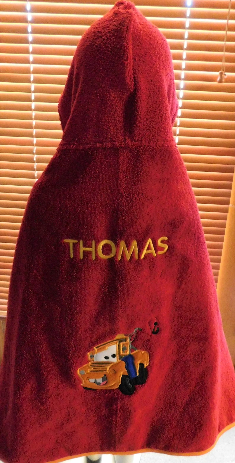 Puede incluir: Toalla con capucha roja con el nombre "THOMAS" bordado en amarillo. Un cami&oacute;n de remolque amarillo con una cara sonriente est&aacute; bordado en la parte inferior de la toalla.