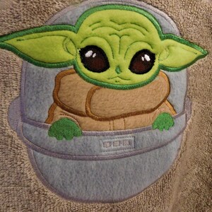 May include: A green and brown embroidered character with large eyes, wearing a brown vest and sitting in a blue spaceship. The character is from the Star Wars franchise.