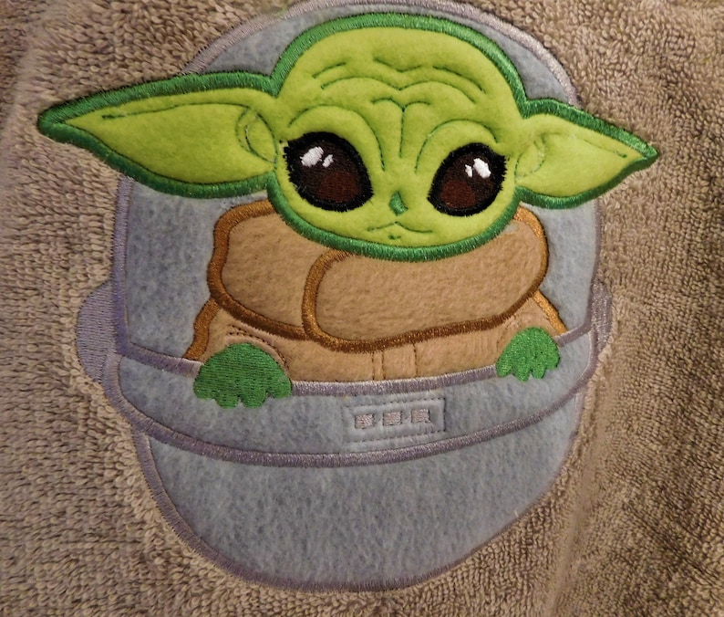 May include: A green and brown embroidered character with large eyes, wearing a brown vest and sitting in a blue spaceship. The character is from the Star Wars franchise.
