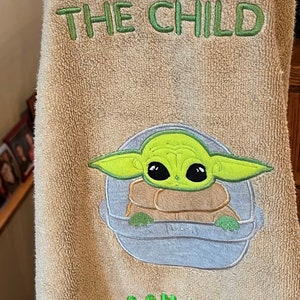 May include: A tan towel with green embroidery that says "Grogu The Child" and "Miller". The embroidery also includes a cartoon image of Grogu, a green alien character from the Star Wars franchise, in a floating pod.