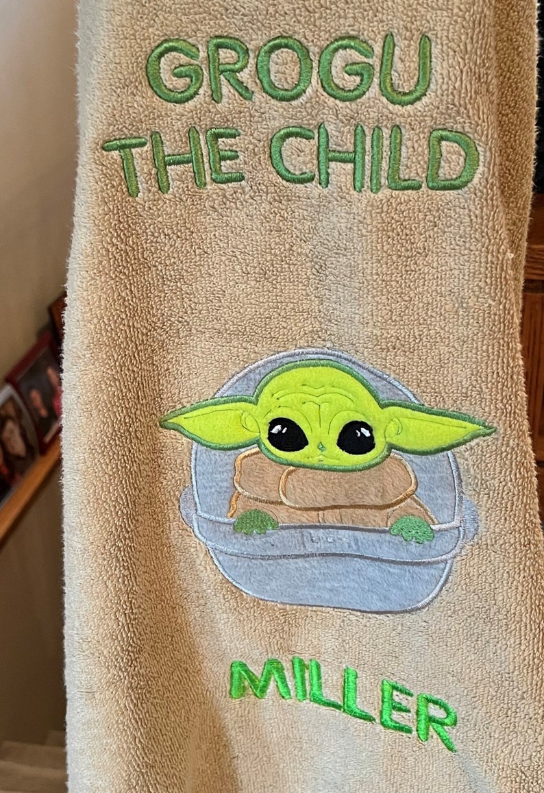 May include: A tan towel with green embroidery that says "Grogu The Child" and "Miller". The embroidery also includes a cartoon image of Grogu, a green alien character from the Star Wars franchise, in a floating pod.