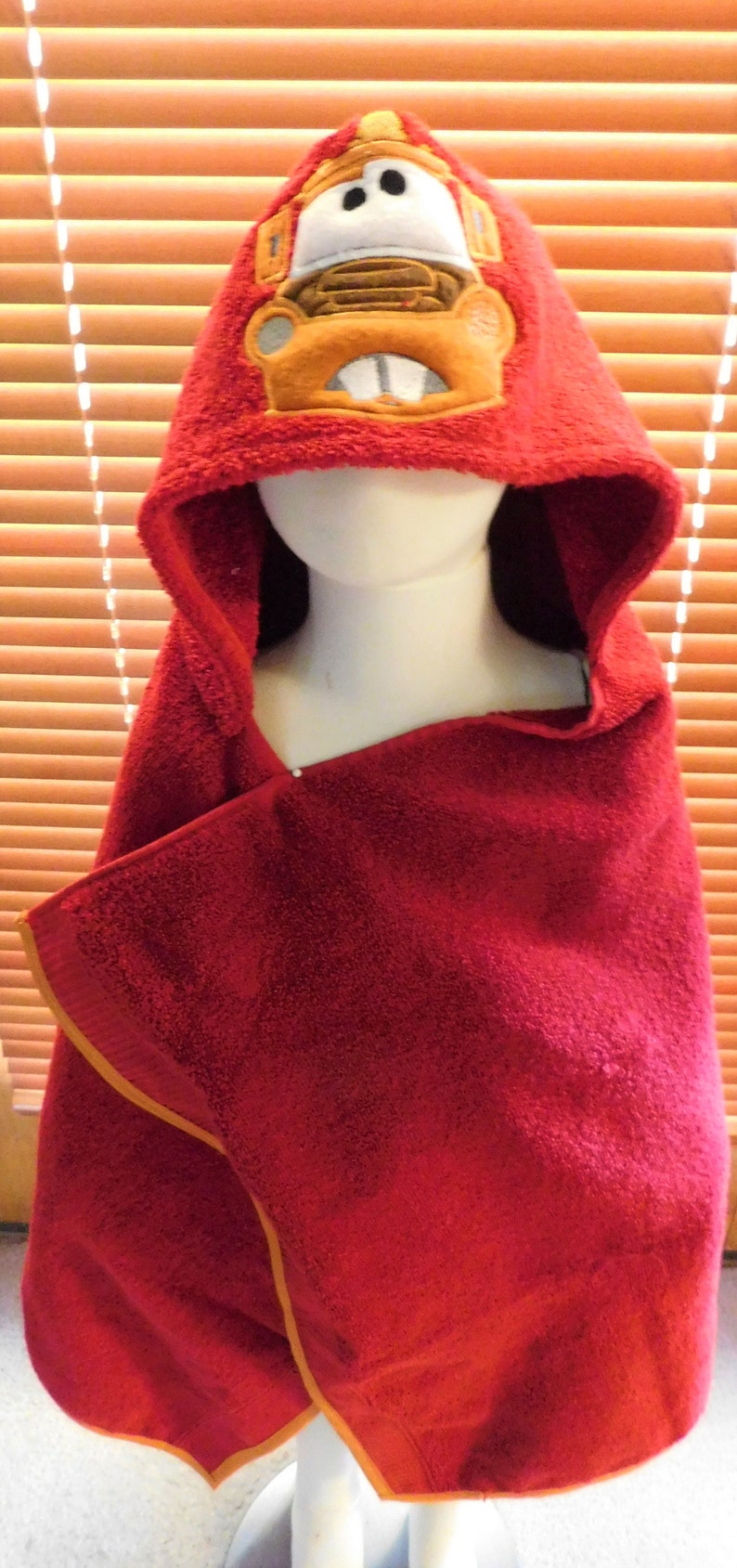 Puede incluir: Toalla con capucha roja con un cami&oacute;n de dibujos animados amarillo y naranja en la capucha. La toalla tiene un ribete amarillo alrededor de los bordes.