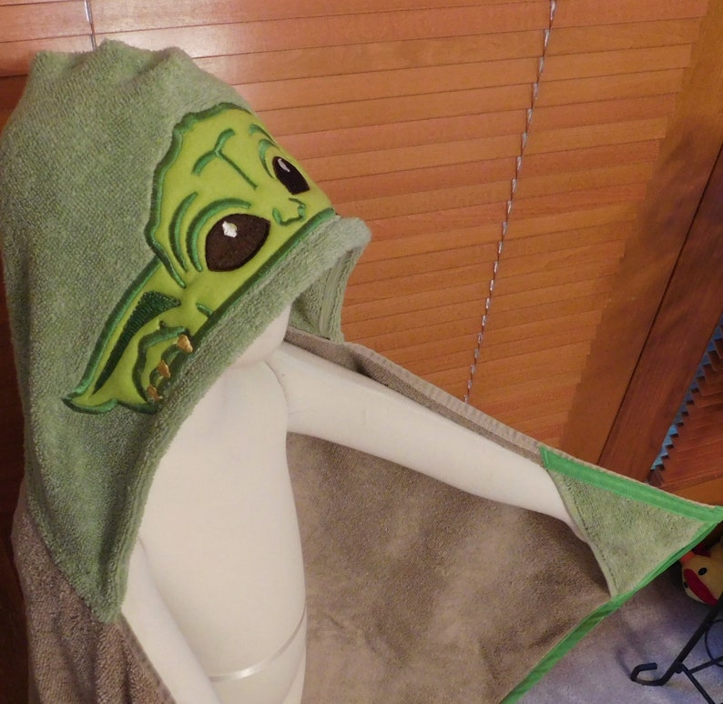 May include: Green hooded towel with an embroidered image of a green alien character with large eyes. The towel is made of soft, absorbent material.