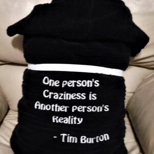 May include: Black fleece throw blanket with white embroidered text that reads "One person's Craziness is Another person's Reality - Tim Burton".