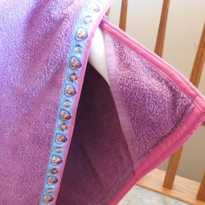 May include: Purple fleece hooded towel with a pink trim and a blue ribbon featuring cartoon characters.