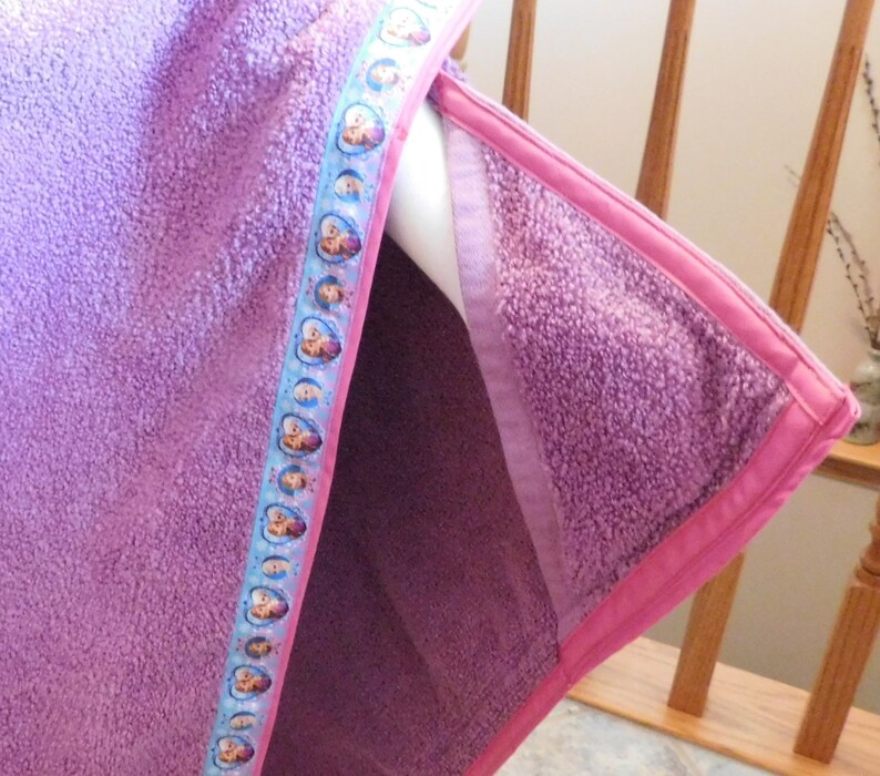 May include: Purple fleece hooded towel with a pink trim and a blue ribbon featuring cartoon characters.