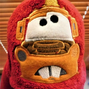 Puede incluir: Sombrero de felpa rojo con una imagen bordada de un coche de dibujos animados en marr&oacute;n, naranja y amarillo con dos ojos grandes.